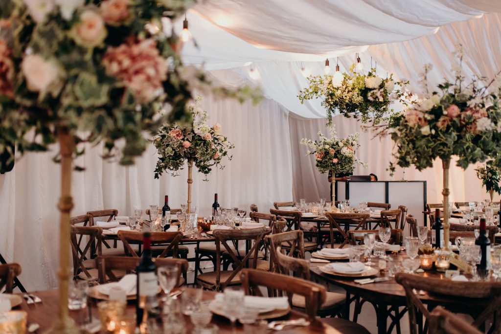Floral arrangements set up at glenfall house in Cheltenham in their marquee, Floral wreaths with white and pink flowers and fresh greenery added hanging from the ceiling,  10ft round tables with a large floral arrangement on gold pedestals as a centrepiece surrounded by coloured tealight holders and place settings of our gold charger plates and classic cutlery and glassware.
