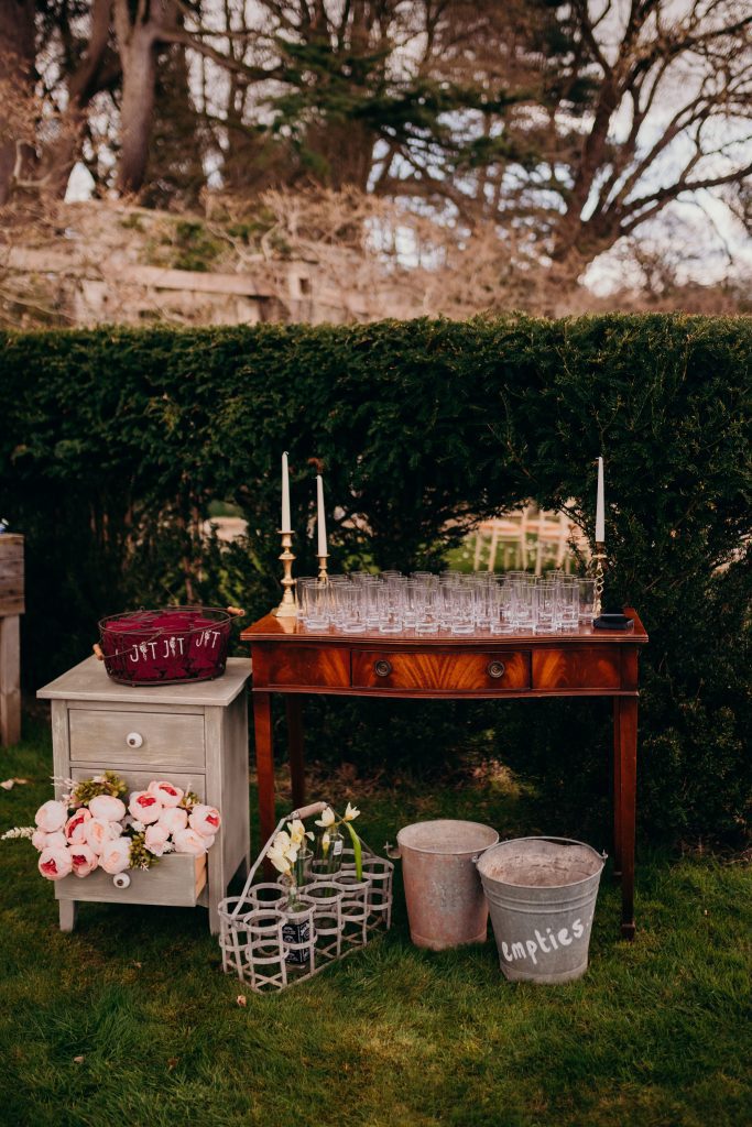vintage wooden console table with three brass candlesticks and white candles with half pint glasses for bottled beer in front of green hedgerow with a small shabby chic bedside table unit with a basket filled with glass holders on top and in front are two rustic tin buckets for all of the empty bottles