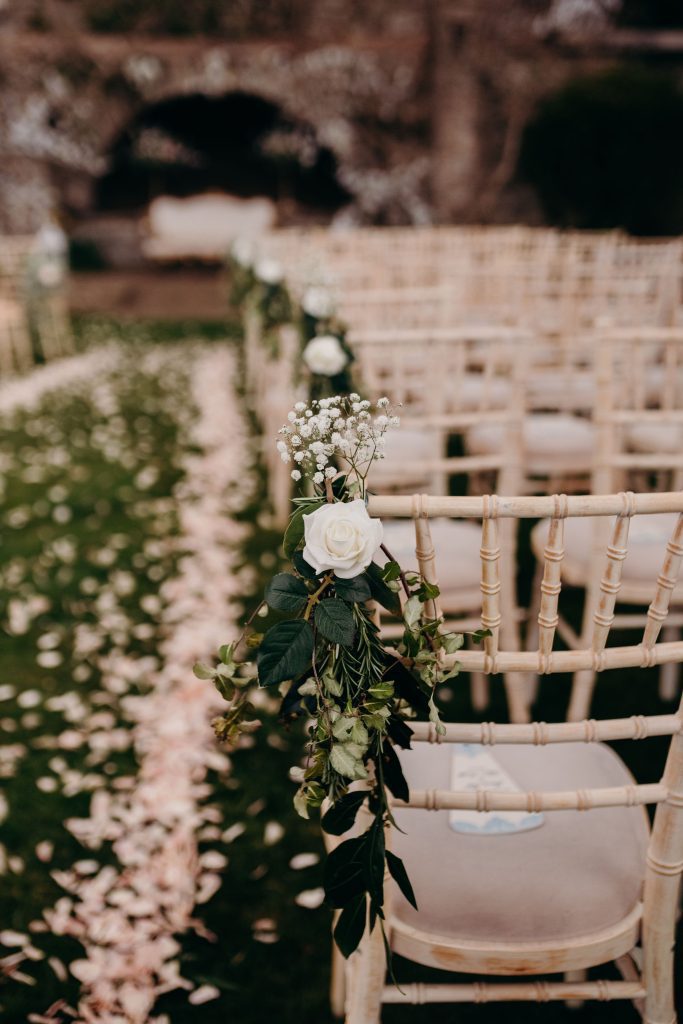 Close up shot of a white rose, fresh greenery and gypsophila tied to the aisle chair with scattered white petals lining the aisle