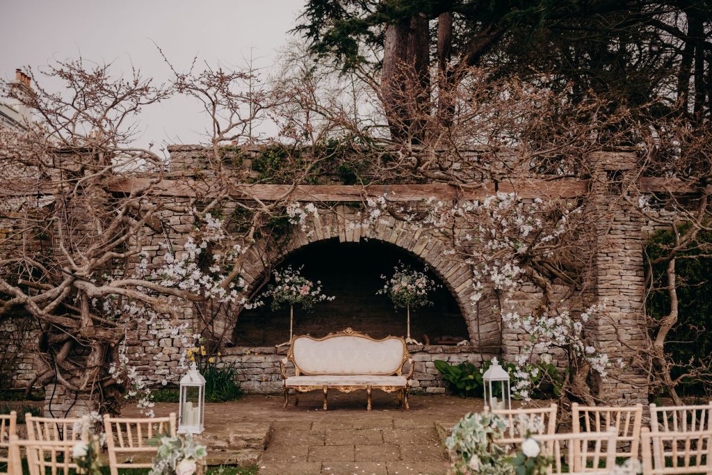 At Glenfall house, Cheltenham, the lower lawn has been set up for a ceremony, with a white and gold vintage sofa in front of a stone archway Blossom branches have been fed into a yet to bloom Wisteria and two pedestal floral arrangements stand either side of the sofa