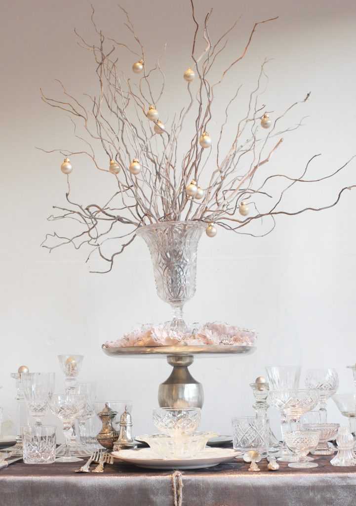 A silver cake stand used as a pedestal for a vintage cut crystal vases filled with twigs of twisted willow and small hanging baubles. the table is set up with vintage crockery and vintage cut crystal glassware