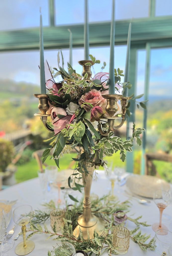 a centrepiece of a gold candelabra with a wreath of faux flowers in dusty rose  and fresh greenery including purple ribbons, around the base is fresh greenery and ivy for hire