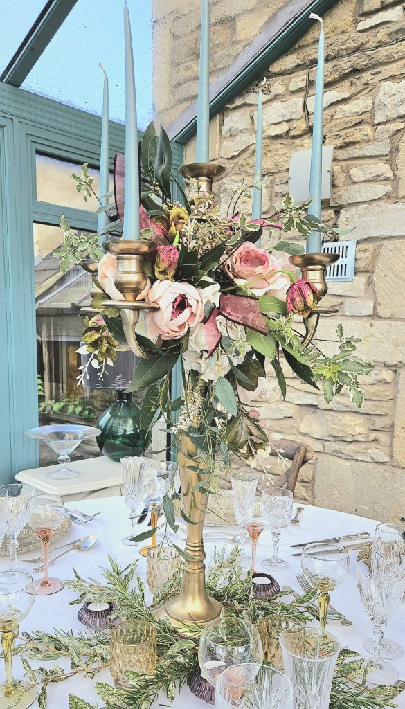 a centrepiece of a gold candelabra with a wreath of faux flowers in dusty rose  and fresh greenery including purple ribbons, around the base is fresh greenery and ivy for hire