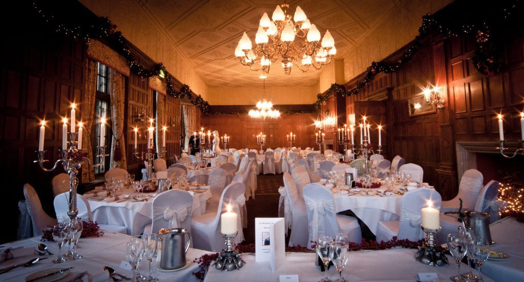 Edwardian panelled dining room set up with round tables covered in white table cloths and white covered chairs with a bow sash and in the centre of each table is a silver candelabra with a wreath of red berries around the base all available to hire