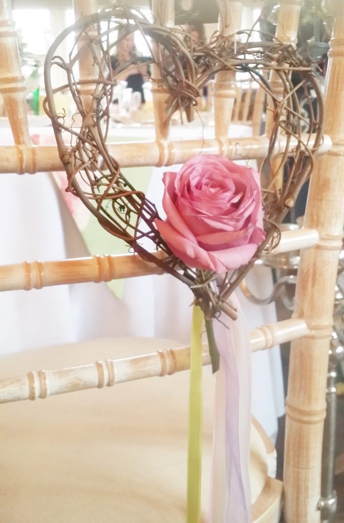 Wicker stick twisted heart with a beautiful pink rose at the base and pale ribbons hanging from the bottom and attached to the back of the chair as a chair hanging for hire