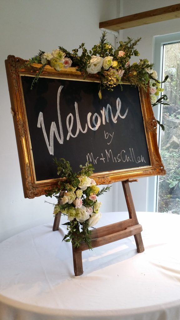 Welcome in white on a blackboard inside a gold picture frame with garlands of flowers round and sat on a wooden easel to hire