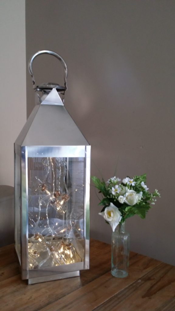 a silver and glass lantern filled with a string of hanging lights with a recycled glass bottle and a posy of flowers in it next to the lantern to hire