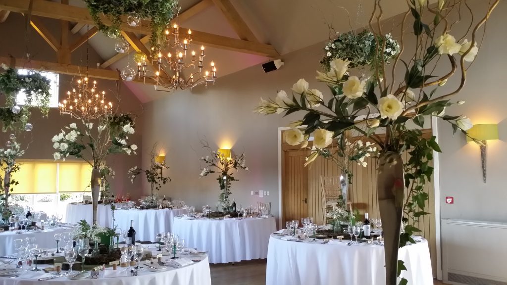 tall white and green wedding table centre decor with lisianthus ivy and twigs, surrounded by a hessian tablecloth and wooden slices as log tealight holders and green stemmed wine glasses at Hyde barn in the Cotswolds available for hire