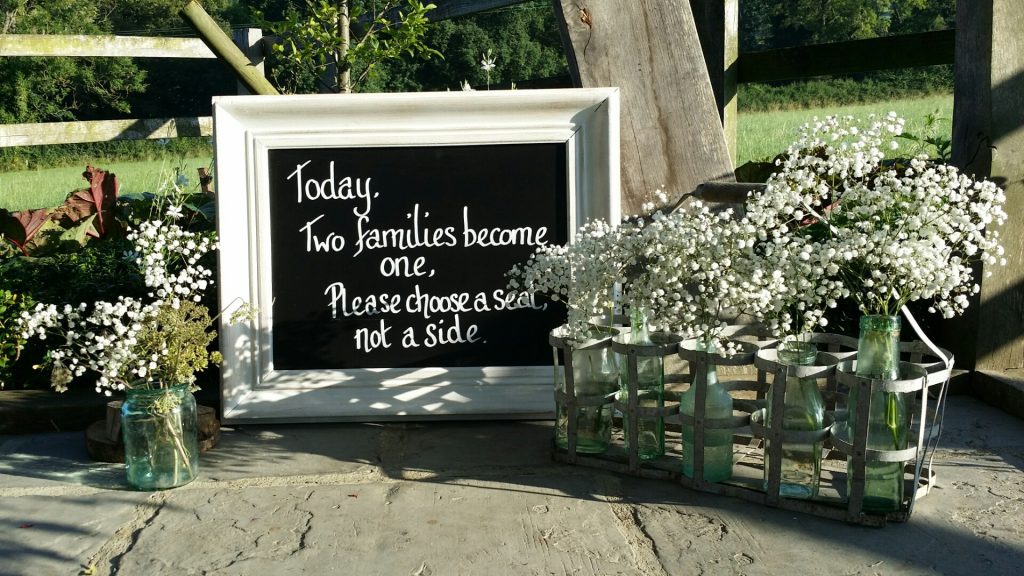 At Hyde barn in the Cotswolds wedding there is a white picture frame with black chalk board and a wedding quote "Today, two families become one, please choose a seat not a side" and recycled glass bottles to each side filled with gypsophila