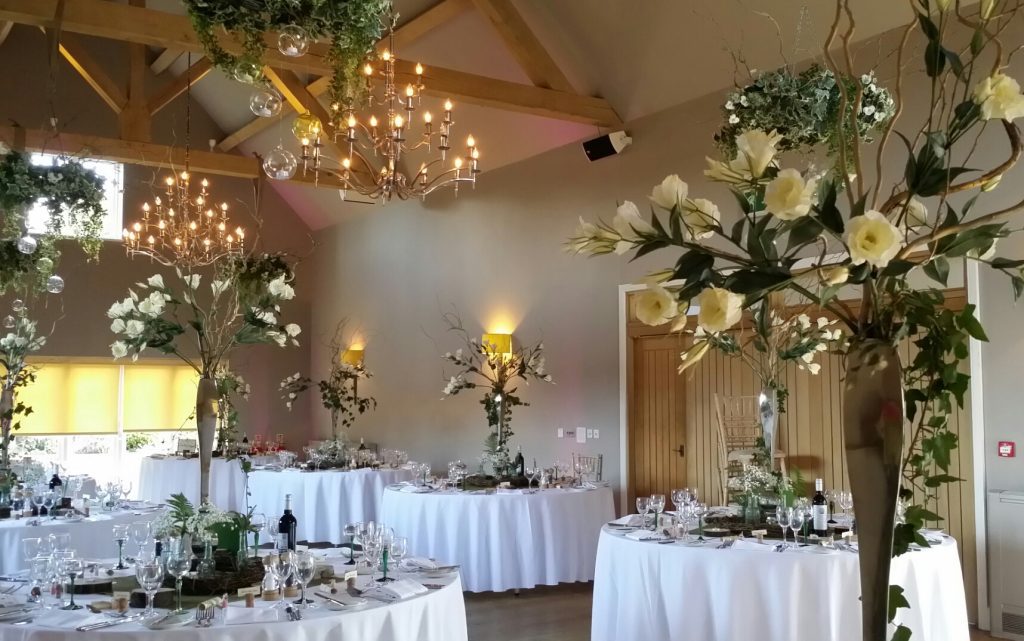 tall white and green wedding table centre decor with lisianthus ivy and twigs, surrounded by a hessian tablecloth and wooden slices as log tealight holders and green stemmed wine glasses at Hyde barn in the Cotswolds available for hire