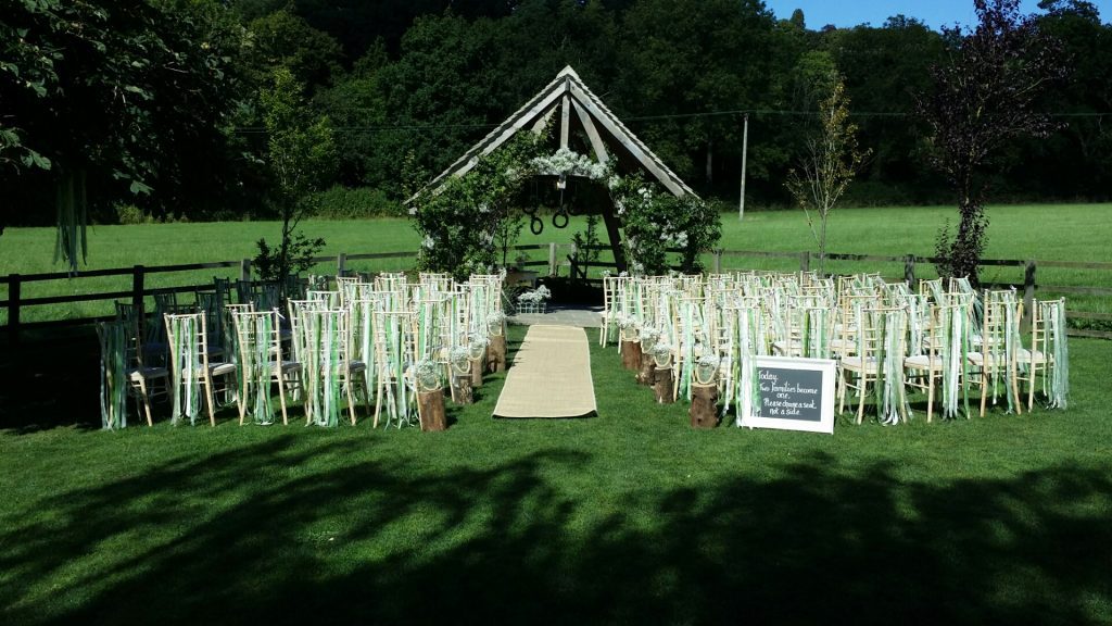At Hyde barn in the Cotswolds Chivari chairs are set up for the wedding ceremony in the garden in front of the pergola each chair has a string of white and different shades of green ribbon tied across the back and a sprig of gypsophila on the aisle side. Also running down the aisle are tree stumps with a glass jar with a rope handle and a large bunch of gypsophila in it.