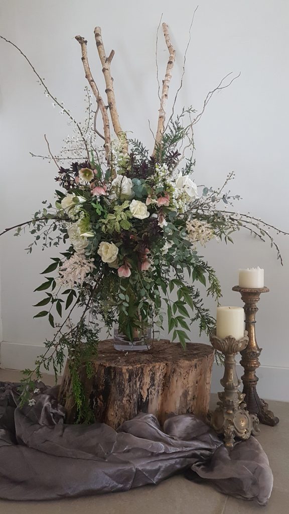 floral arrangement in a vintage vase sat on a tree stump with two gold Blenheim ornate candlesticks with pillar candles next to it for hire