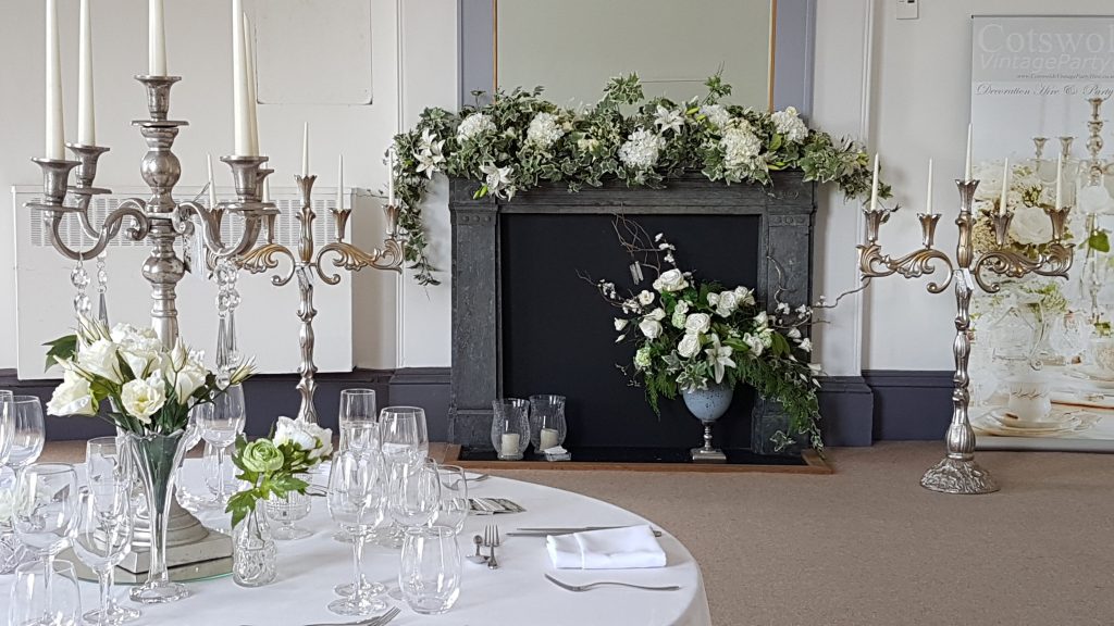 wedding fayre set up with a table to the left and a silver candelabra as the centrepiece and behind is a fire place with a faux flower garland in white and green and two large ornate candelabras one on each side for hire