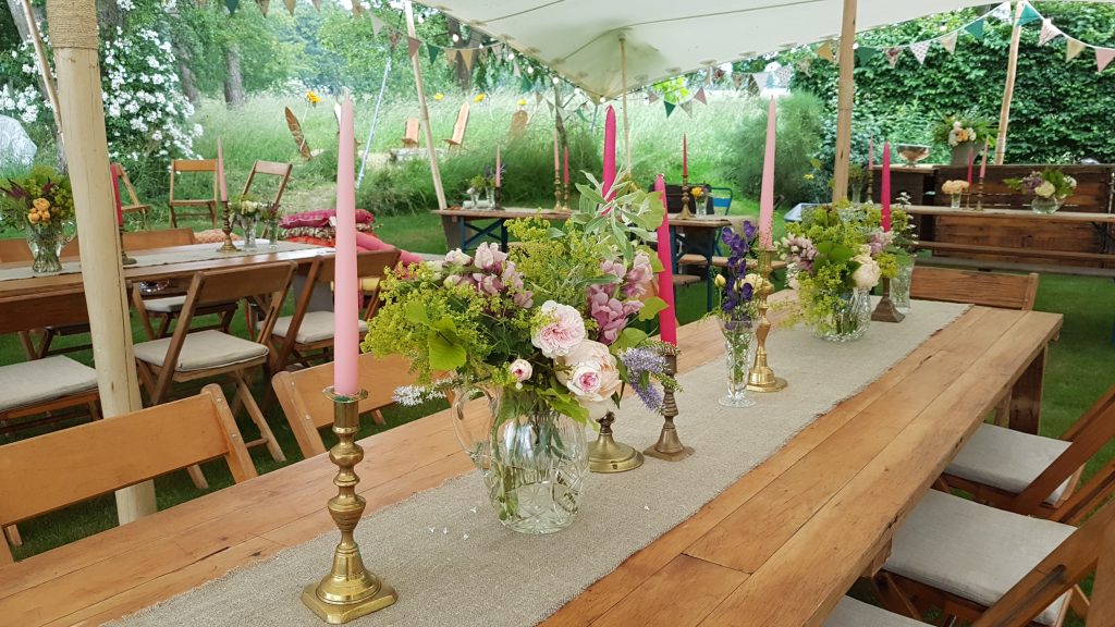 under a stretch marquee with garden party bunting wrapping the outside, there are many wooden trestle tables with a hessian runner down the middle of each and on top are a mix of brass candlesticks with different shades of pink candles an vintage cut crystal glass vases and jugs fill with a variety on wild flowers and greenery