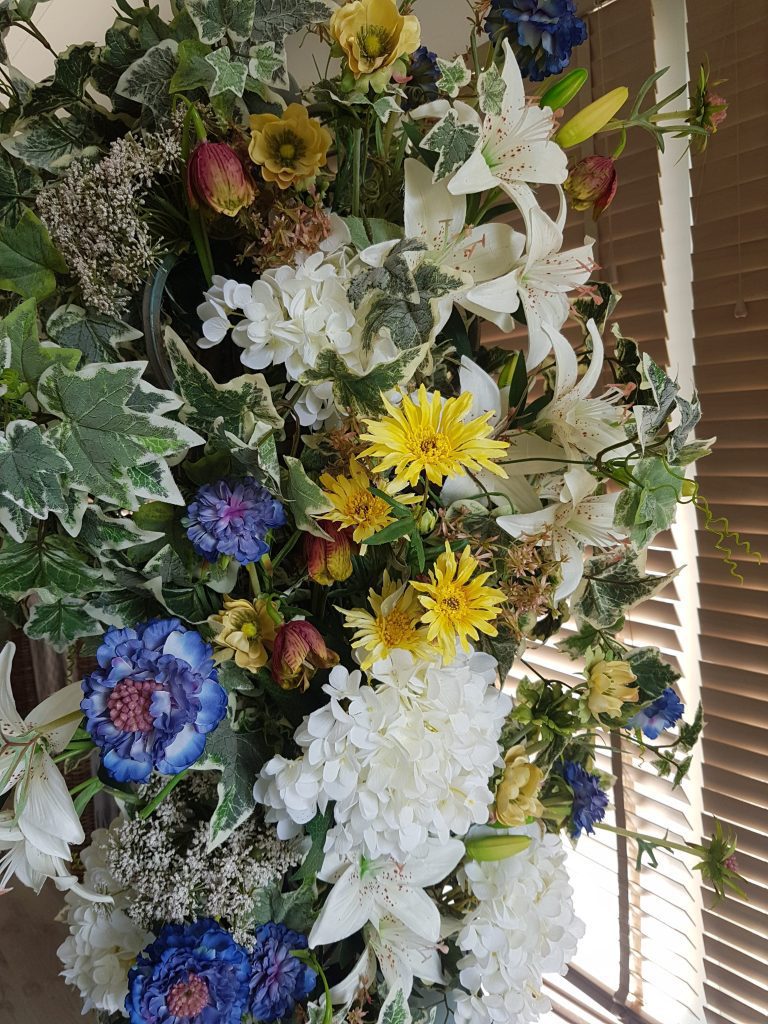 A pillar of fake flowers and greenery with green ivy, white lilies and White hydrangeas, Blue cornflowers, and yellow anemones and gerberas.