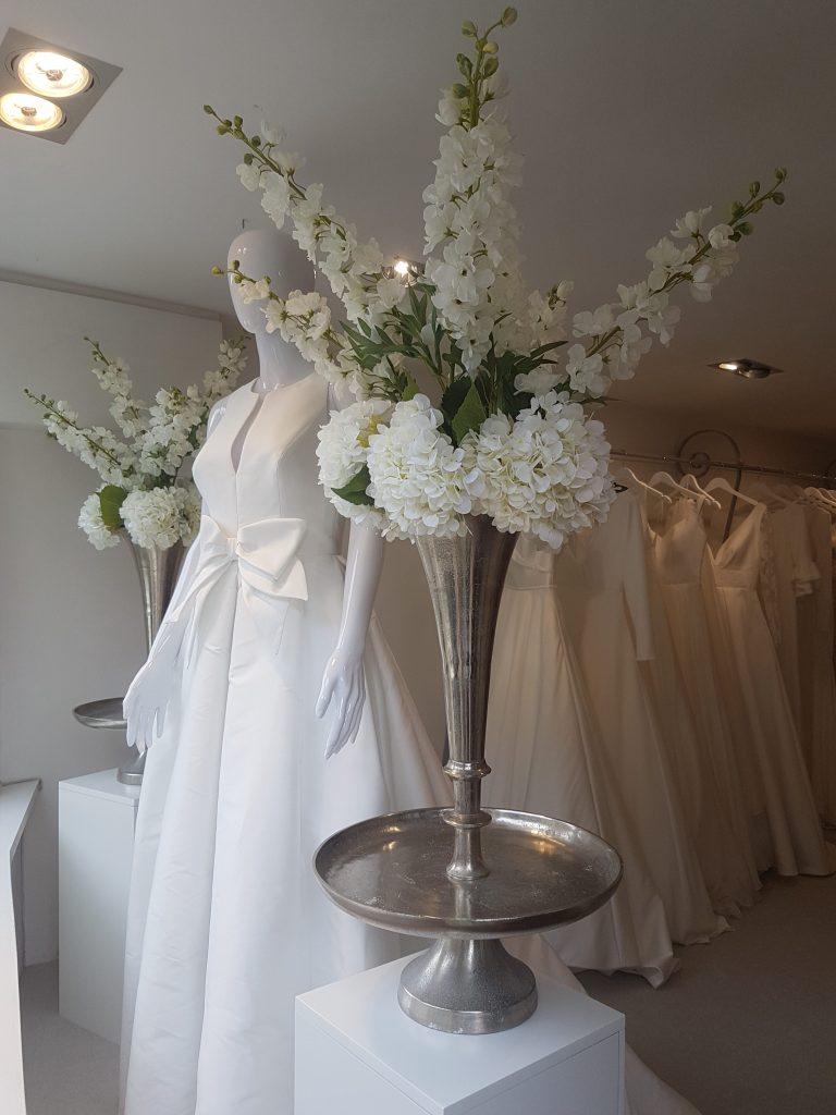 At the white room in Minchinhampton either side of a manikin in a wedding dress is a large silver trumpet vase with fake white hydrangeas and lisianthus for hire