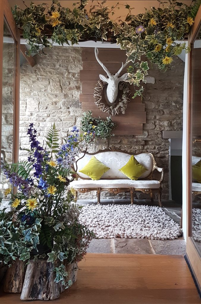 Faux flower garland above a double door was with yellows and blues added in to the arrangement, to the left is a fake floral arrangement matching the garland on a large tree stump in the room behind is a vintage white chair with a gold frame, below a white stags head for hire