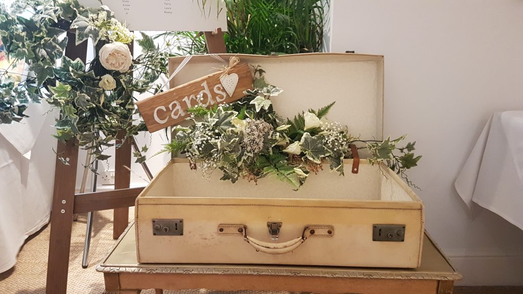 A flower arrangement of white and pink roses, light pink peonies, white hydrangeas with gypsophila, filled with soft green ivy and green ferns in a vintage cream suitcase for wedding cards and gifts