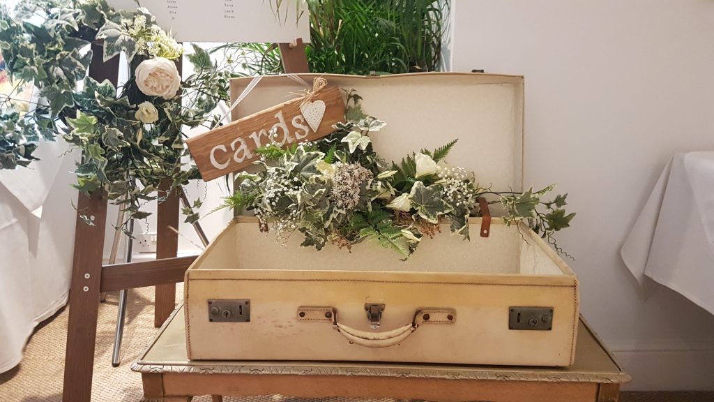 A flower arrangement of white and pink roses, light pink peonies, white hydrangeas with gypsophila, filled with soft green ivy and green ferns in a vintage cream suitcase for wedding cards and gifts