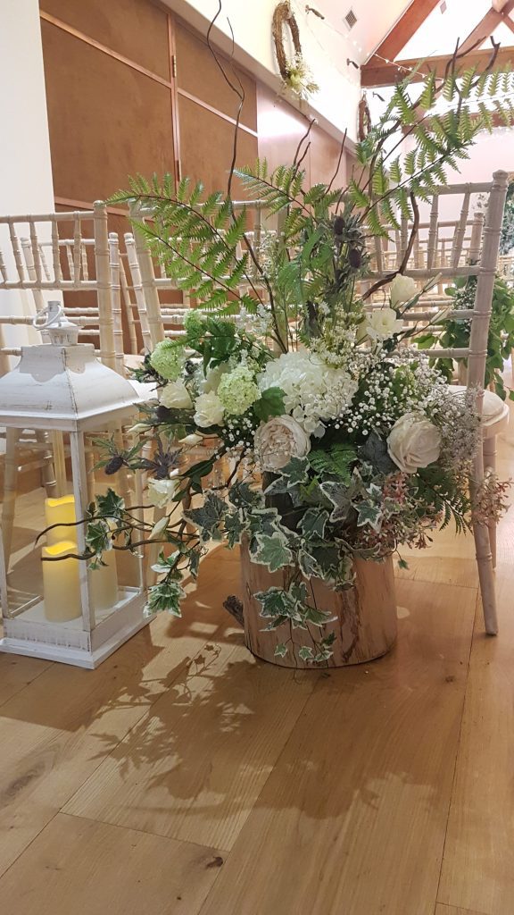 A flower arrangement of white and pink roses, light pink peonies, white hydrangeas with gypsophila, filled with soft green ivy, ferns and twisted willow on a tree trunk, next to this is a large white lantern.