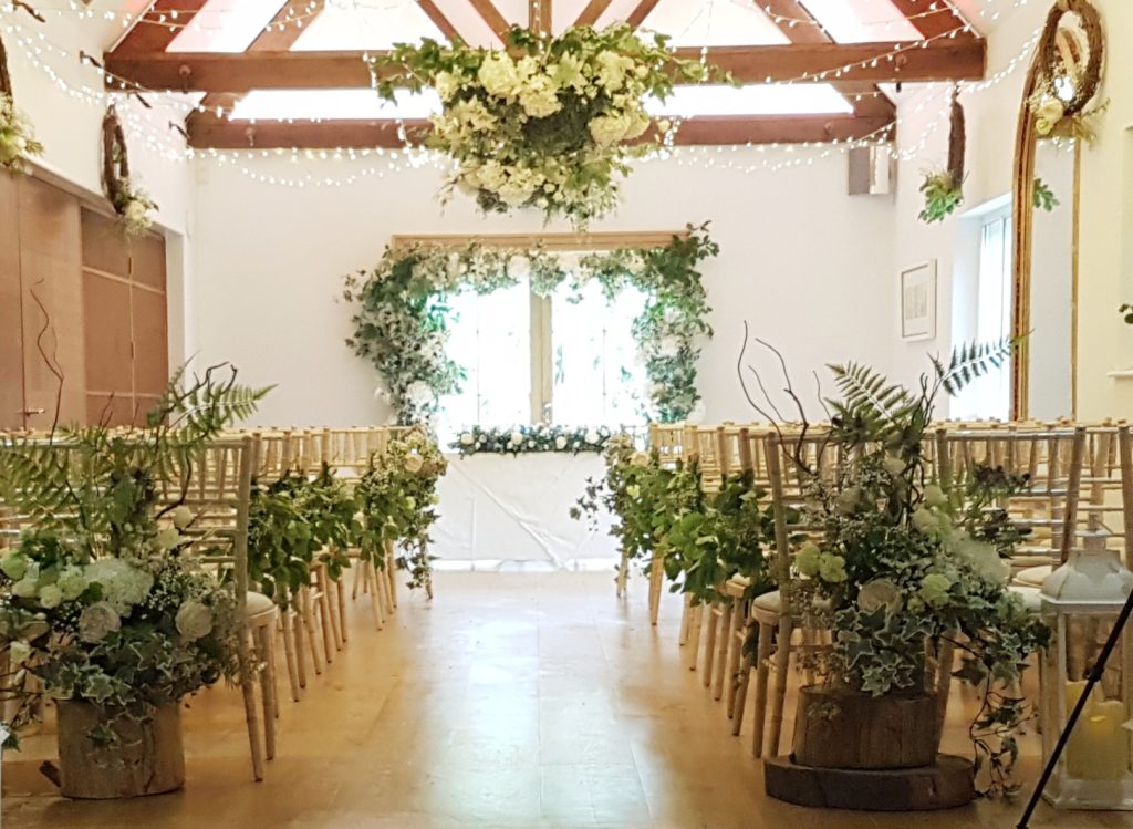 The Cotswold hotel in Chippen Camden Set up for a ceremony with a flower arrangement at the back of each aisle chair and fresh greenery attached to each of the chairs. hanging from the wooden beams are white and green floral wreaths and fairy lights at the end of the aisle is a beautiful flower arch framing the window behind where the newlyweds will sit