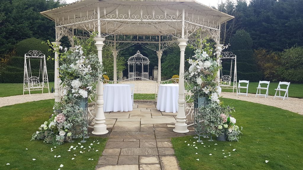 At Manor by the lake in Cheltenham, the Cotswolds, we set up the ceremony under the while ornate pergola, either side of the entrance are two pillars covered with quality fake flowers such as roses, hydrangeas and lilies and greenery.