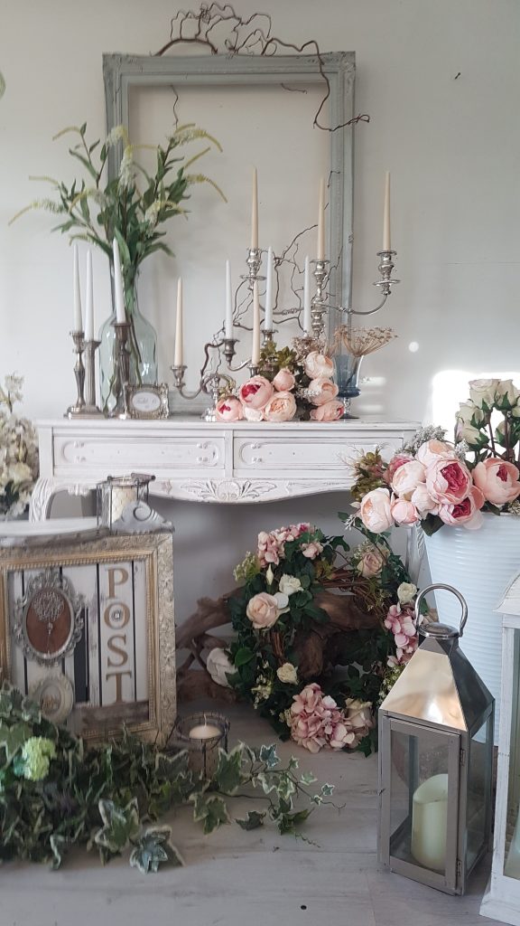 vintage silver beauty and the beast style chandeliers and Blenheim gold pillar candle sticks with coral peonies and dried flowers in front of a duck egg blue picture frame on the floor is a white vintage post box, silver lantern and a white bucket of faux flowers for hire