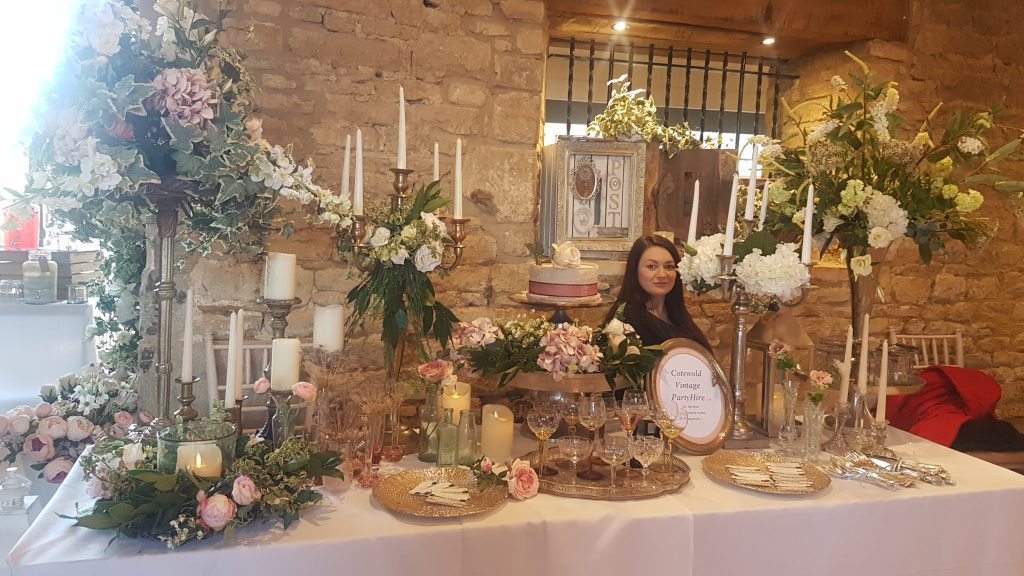 wedding fair set up at the great tythe barn in Tetbury, with gold an silver candelabras, glass, brass and ornate candlesticks, large floral arrangements, gold charger plates, large two tier silver cake stand vintage cut crystal vases and vintage cut crystal vases for hire