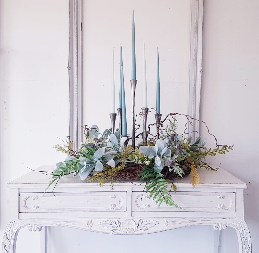 Seven stick candelabra with each spoke at a different height in silver with blue and white candlesticks and a wreath of green foliage around the base and a White vintage picture frame framing it to hire