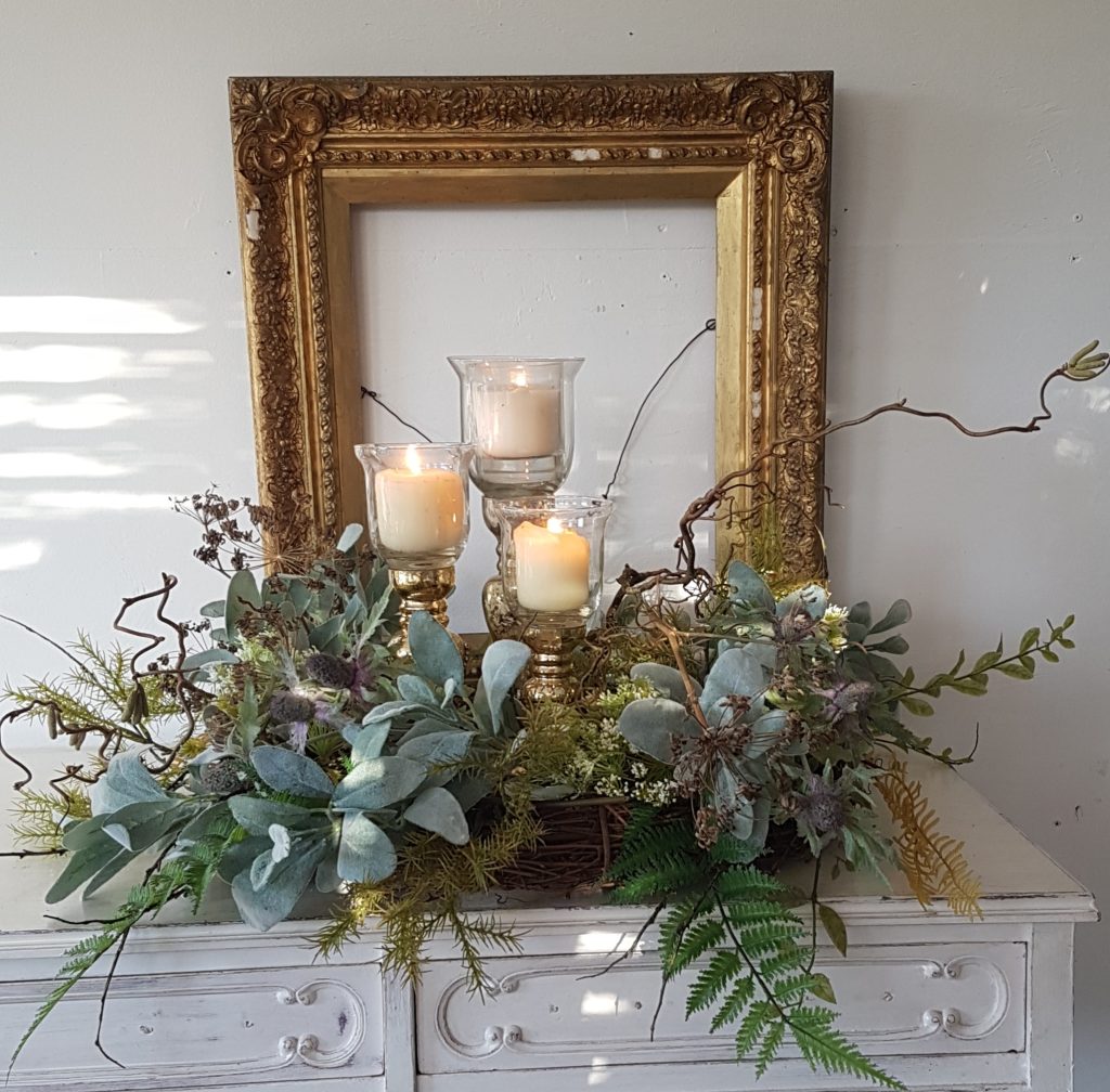 Vintage gold picture frame with glass gold candle holders at different heights, with a wreath around the base and green foliage and ferns, sage, bay and twisted willow