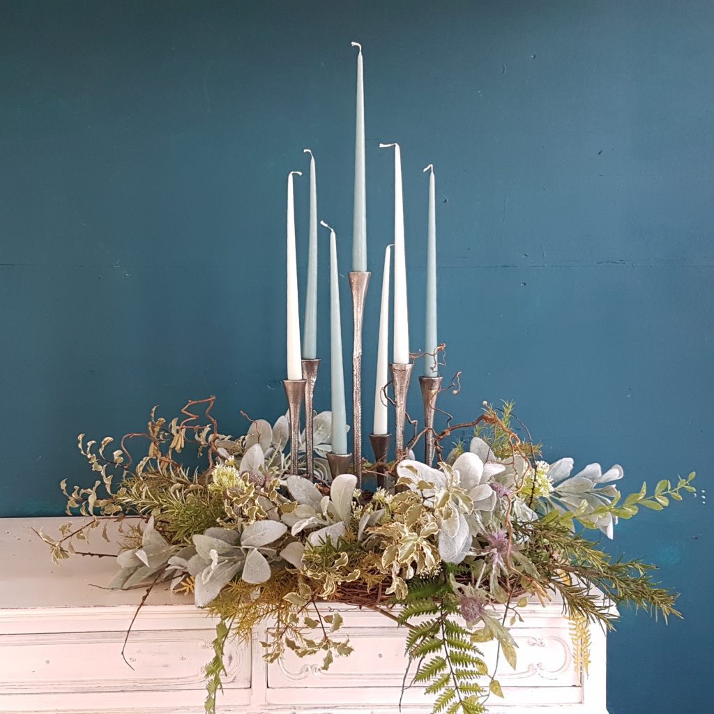 Seven stick candelabra with each spoke at a different height in silver with blue and white candlesticks and a wreath of green foliage around the base and a blue background