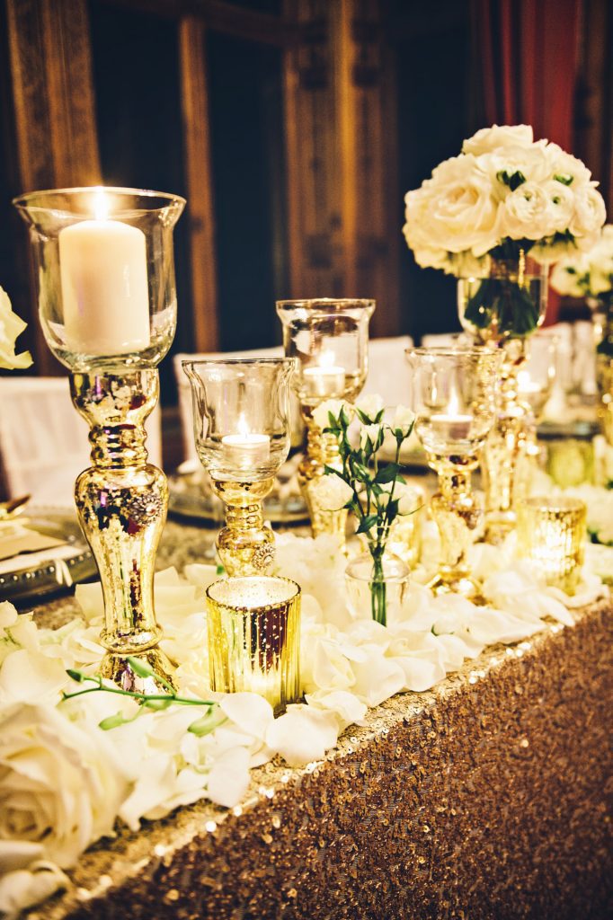 Mercury gold goblet style vase and candleholders and mini tealight holders amongst White rose petals at Manor by the lake wedding venue