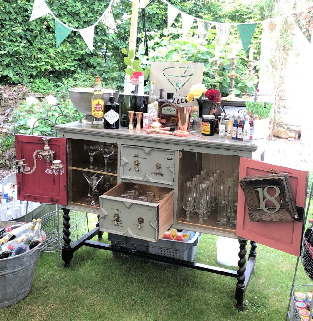 Vintage shabby chic side board with drawers and cupboards turned into a cocktail bar painted in a light grey and sanded back with drinks and glasses on top along with cocktail shakers, mixers and an ice bucket with an ice bucket for mixers also in front on the floor
