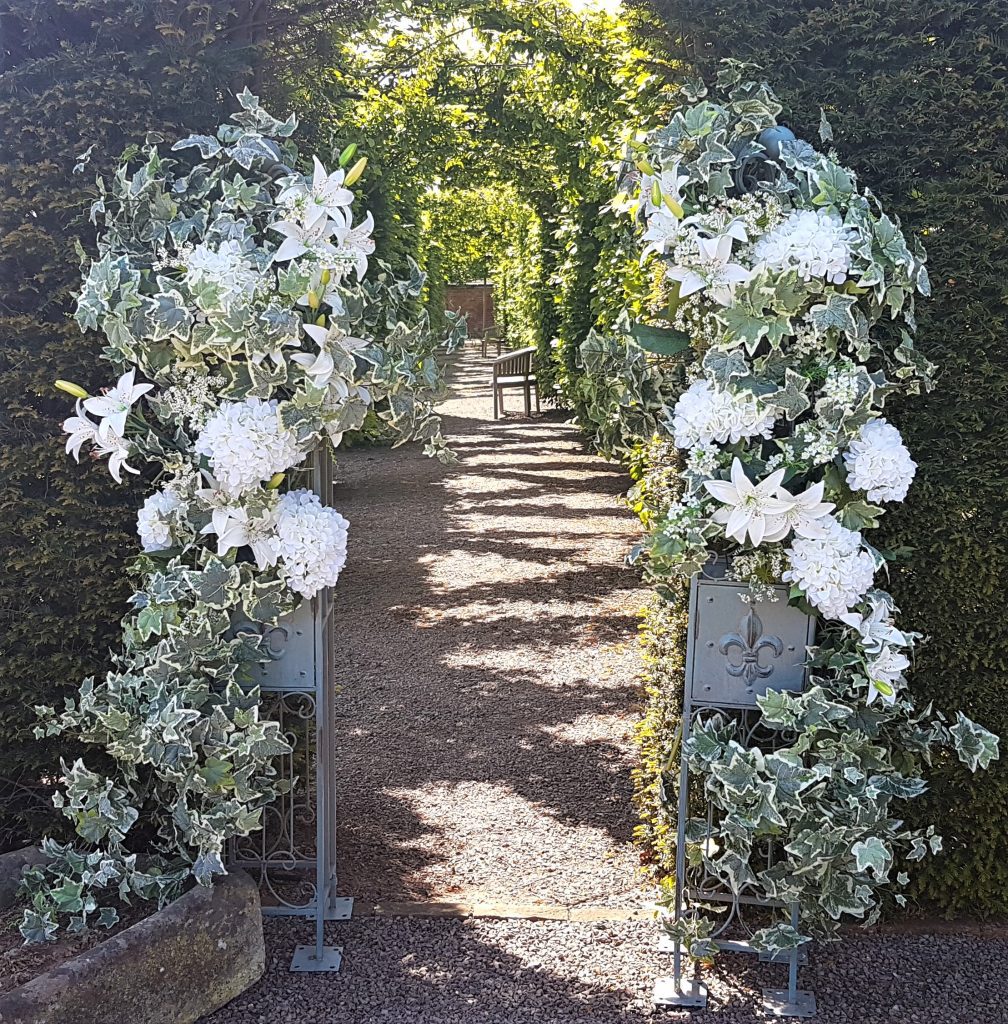 A hedgerow opening and either side of the entrance are two pillars covered with quality fake flowers such as roses, hydrangeas and lilies and greenery.
