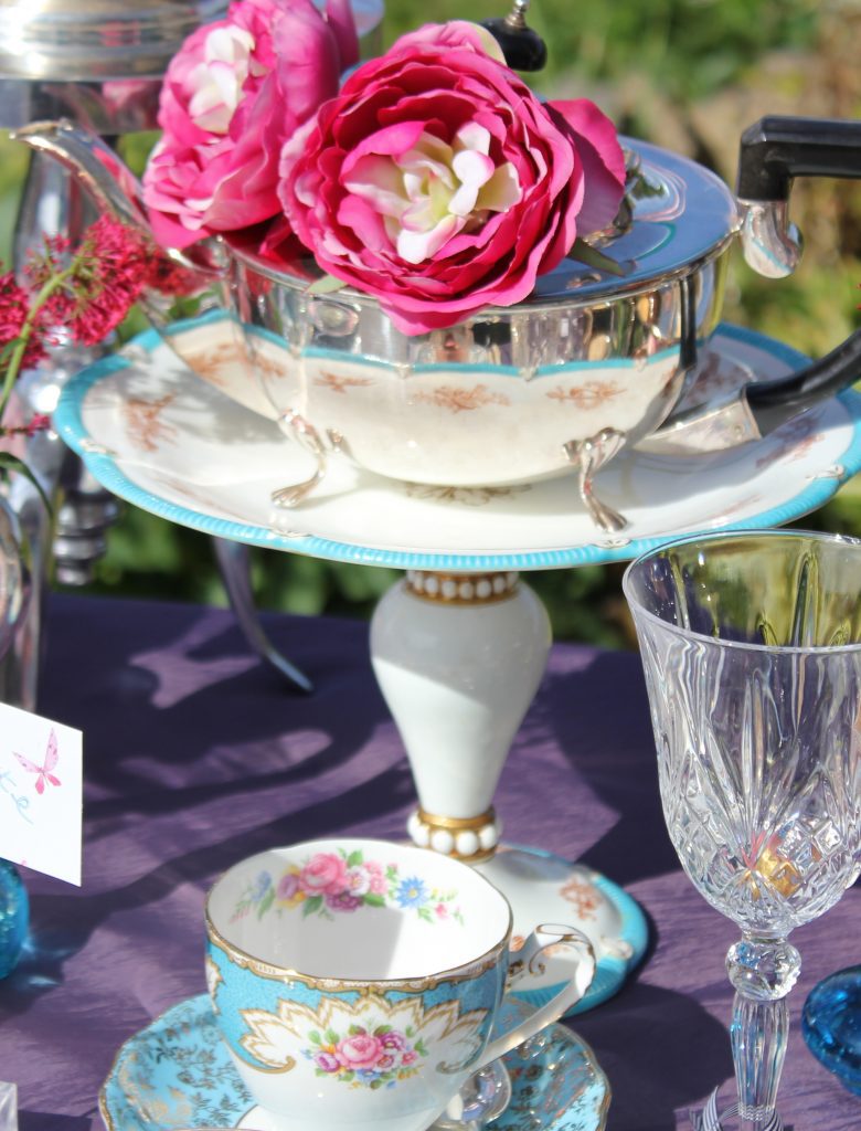 A vintage white china pedestal cake stand with gold detail and a blue trim, on top a silver vintage teapot and in front is a blue and white vintage tea cup and saucer and a cut crystal wine glass