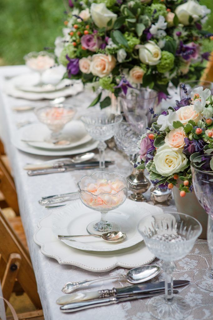 Close up of three vintage tableware set ups of white charger plates then a side plate on top and a vintage glass posy vase on top and silver vintage cutlery of starter main and desserts set t each side and cut crystal wine and champagne glasses and saucers