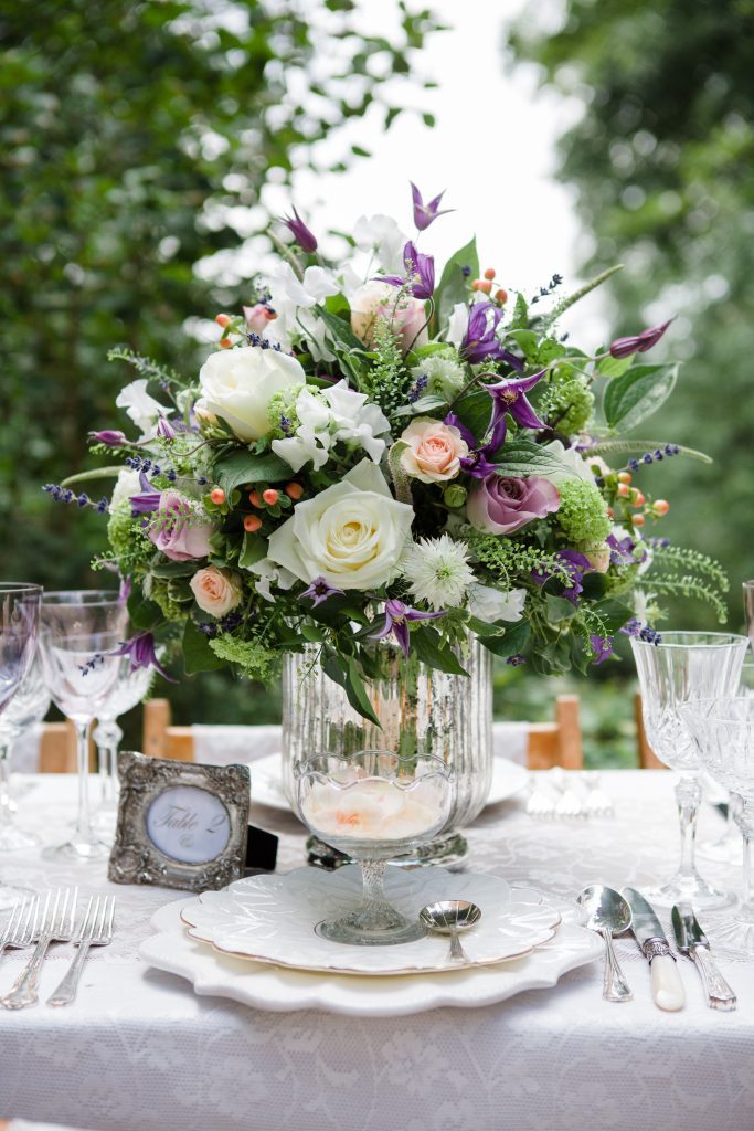 Close up of three vintage tableware set ups of white charger plates then a side plate on top and a vintage glass posy vase on top and silver vintage cutlery of starter main and desserts set t each side and cut crystal wine and champagne glasses and saucers with a silver vintage frame for the table number and a vintage vase filled with a bouquet of stunning white and lilac roses and fresh greenery.