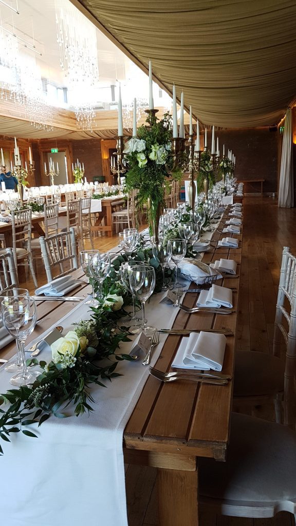 Elmore court in the gilly flower, set up with four rows of trestle with a hessian runner down the middle of each table and filled with gold candelabras with faux flower white and green wreaths and fresh ivy running between each candelabra