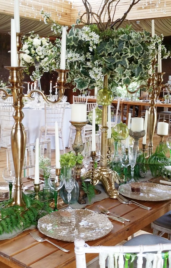 Gold candelabra and gold charger plates with a green ivy and twisted willow tall foliage table centrepiece, brass candle sticks and ornate pillar candle holders on a trestle table with a hessian runner to hire