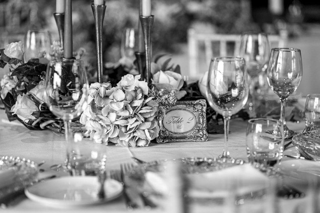 Black and white image of the moulded glass charger plates as a close up with classic silver cutlery and wine and water glasses with a floral wreath around a silver seven stick candle holder
