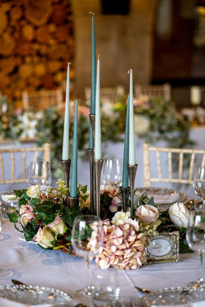 Seven stick candelabra with each spoke at a different height in silver with blue and white candlesticks and a wreath of green foliage and faux flowers around the base with turquoise and pale green candlesticks for hire