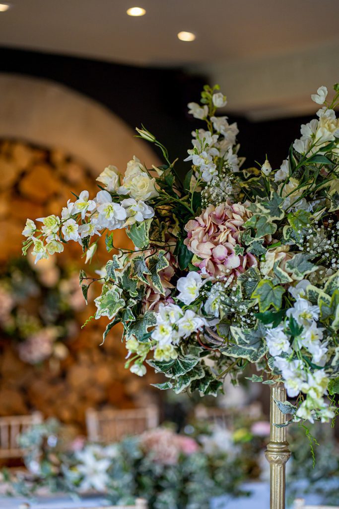 Floral wedding at the bear of Rodborough, a large floral arrangement with fake ivy, lisianthus, pink hydrangeas and gypsophila on a gold pedestal as a centrepiece surrounded by coloured tealight holders with a blurred background