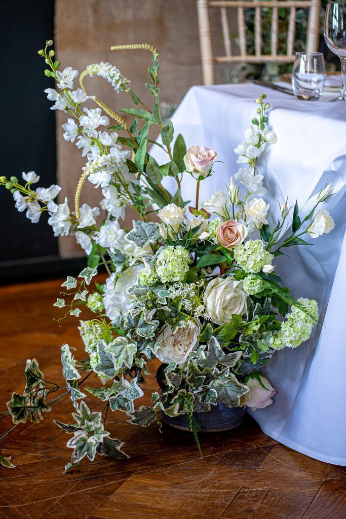 at the foot of the top table is a faux floral arrangement of pink roses, green and white ivy, green ferns, off white hydrangeas and white lisianthus in a light blue jug