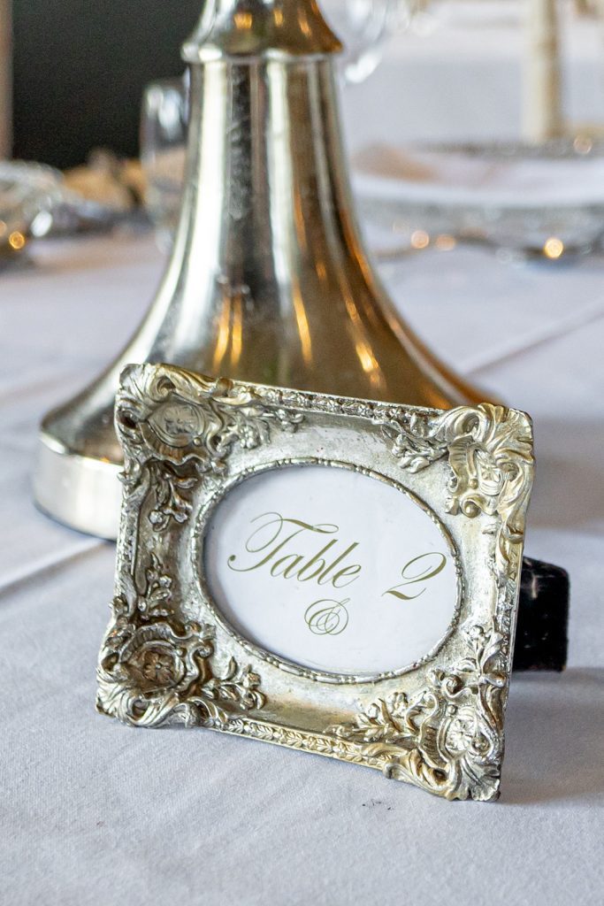 Table name or number in a small silver vintage looking ornate picture frame in front of the base of the silver trumpet vase