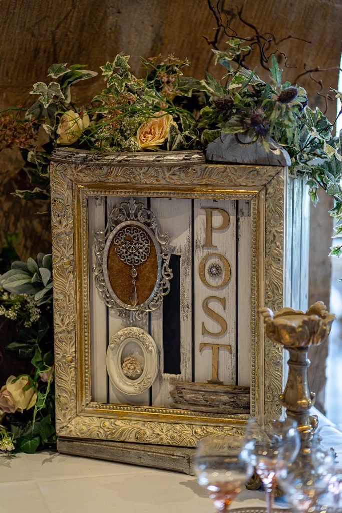 White and gold vintage post box made from a crate painted white with post in gold letters to the right of the opening and two vintage picture frames to the left, covered in fake ivy and faux roses and cow parsley
