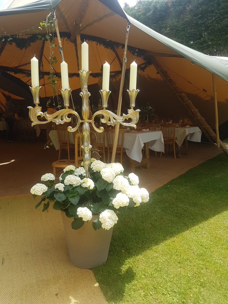 Stunning ornate extra large candelabra with pillar candles in front of a stretch tent marquee with a large plant pot of white hydrangeas in front