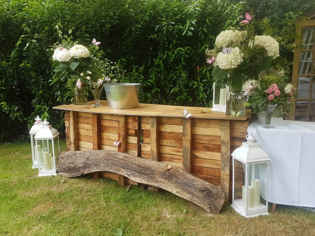 Rustic wooden pop up bar with our rustic silver ice bucket to the left hand side and crystal vases filled with roses , hydrangeas and fresh greenery on either end and on the floor in front are tall white lanterns