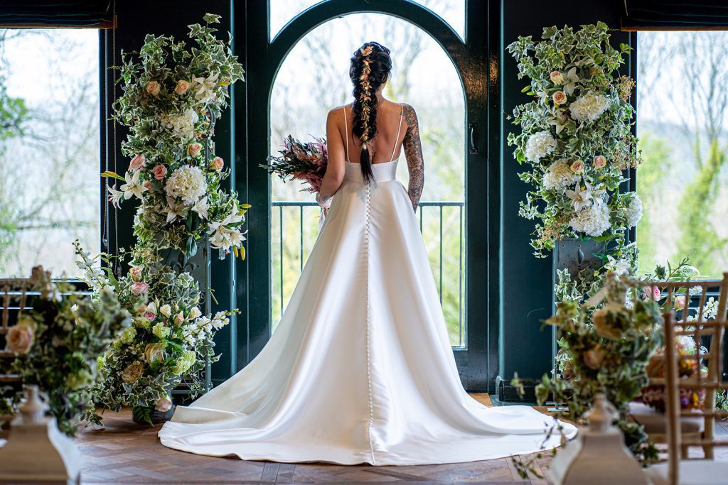 The dining room at the bear of Rodborough set up for a wedding ceremony with two pillars decorated with fake white and green ivy, white hydrangeas and lilies and pink and white roses either side of the window with a jug t the base of each pillar filled with a floral arrangement. in front of the window is a brunette bride with her hair in a fish tail with flowers running down the middle, wearing a timeless satin dress with buttons running down the black to the end of the dress. up the aisle on the end of the chairs are a bunch of flowers to make the pillars and two white lanterns