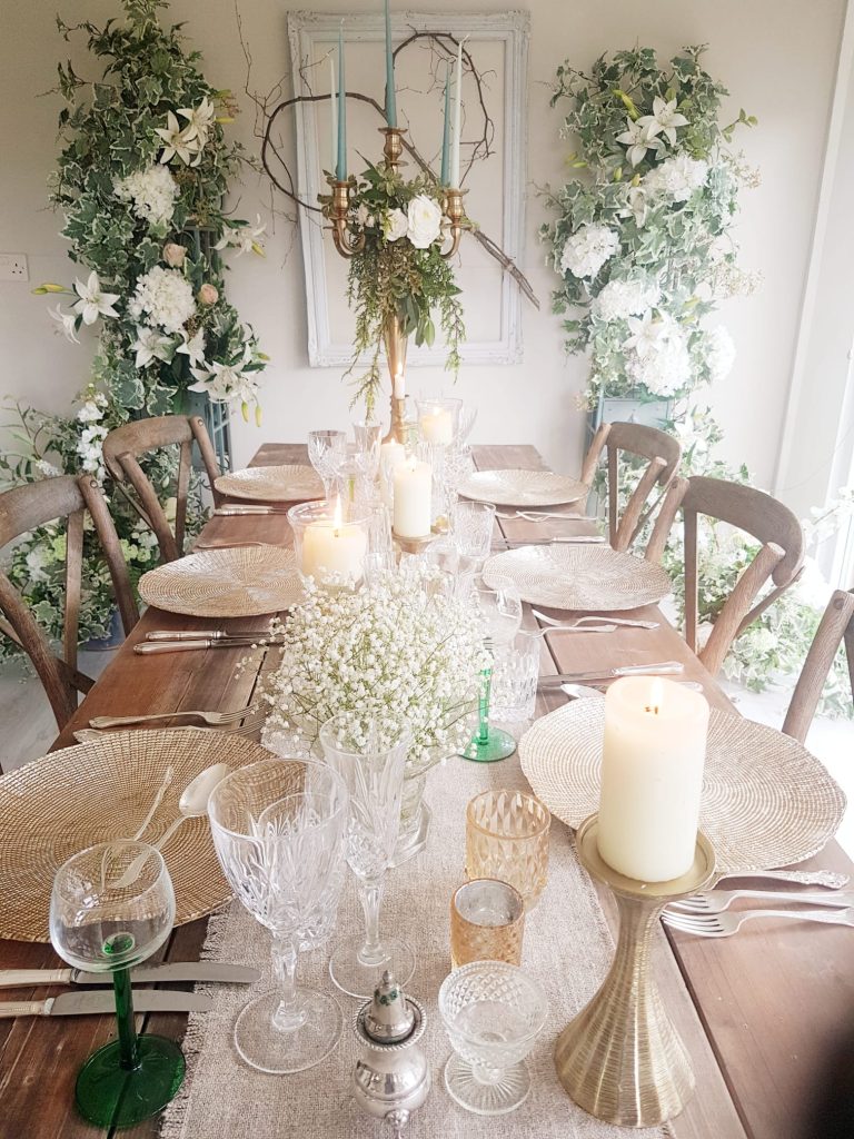 rustic wooden trestle table setup with gold charger plates, gold candelabra decorated with a wreath of white roses and fresh greenery, with place settings made up of vintage cut crystal wine glasses, tumblers and champagne saucers and cut crystal vintage vases with a backdrop of two pillars covered in faux white flowers and green ivy either side of a large vintage photo frame