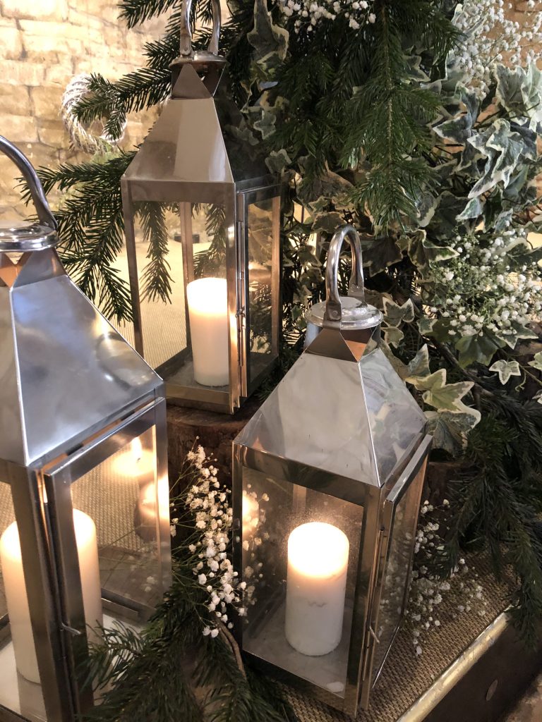 Three silver lanterns on wooden tree stumps in front of a frame of ivy, pine branches and gypsophila, for wedding ideas at great tythe barn, available to hire for weddings and events